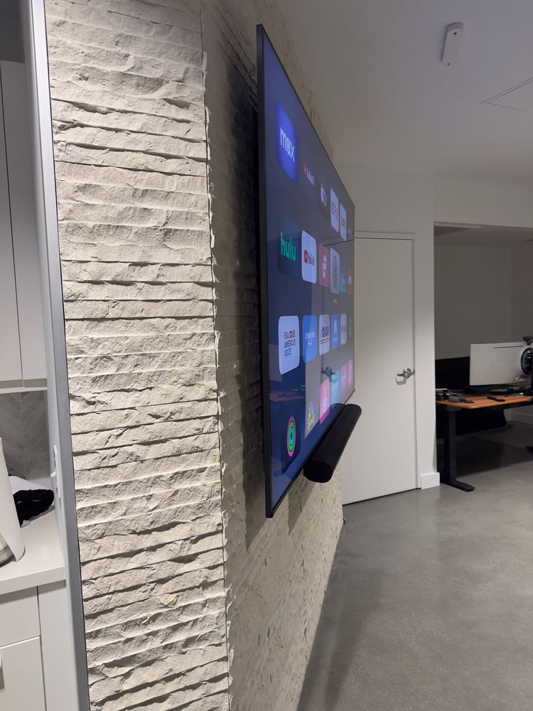 Wall-mounted TV in a modern office hallway beside a white brick wall, with desks visible in the background