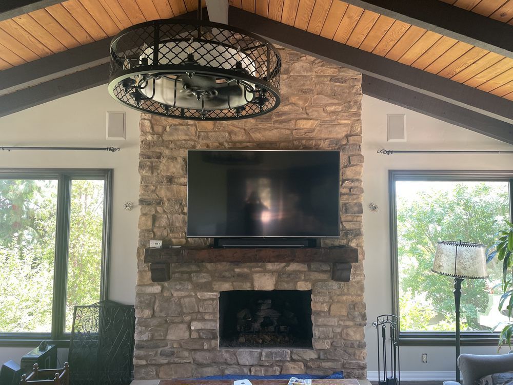 Living room stone fireplace with mounted TV and large windows, beneath a vaulted wood ceiling