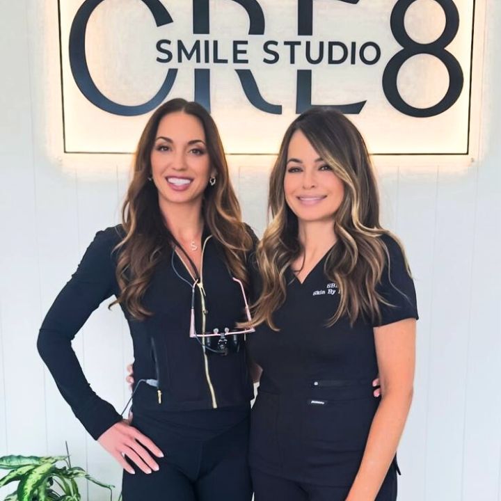 Two women in black outfits standing in front of a Smile Studio 18 sign, smiling at the camera.