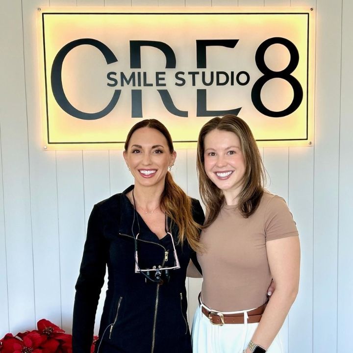 Two women standing smiling in front of a “Smile Studio CR8” sign in a bright office.