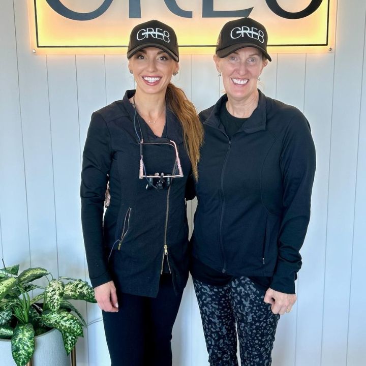 Two smiling people in black outfits and caps standing indoors in front of a sign.