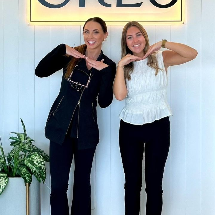 Two people standing indoors, smiling and posing with hands under their chins in front of a sign.