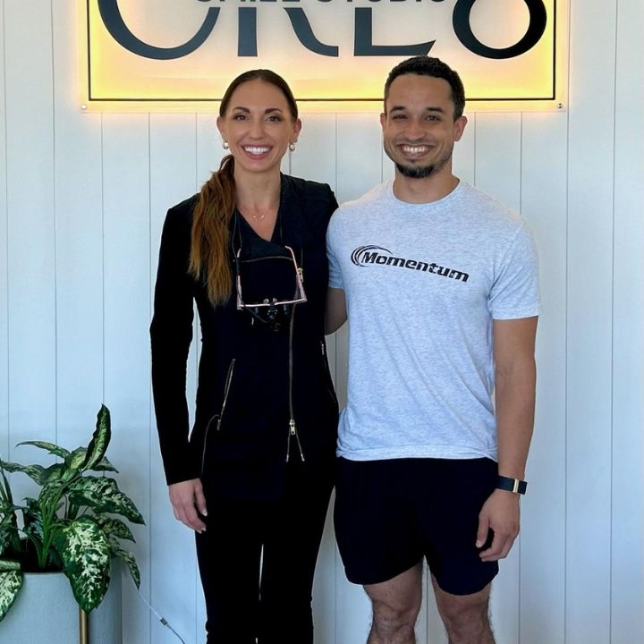 Two people standing indoors in front of a lit sign, smiling at the camera.