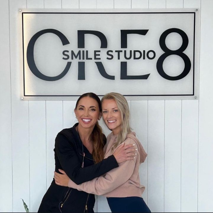 Two women smiling and hugging in front of a CRE8 Smile Studio sign