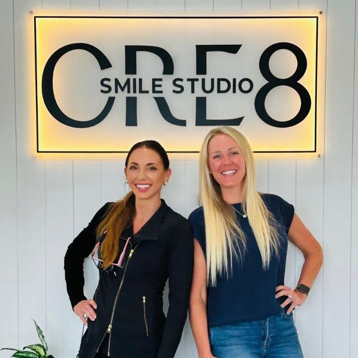 Two women standing in front of a CRE8 Smile Studio sign, smiling indoors.