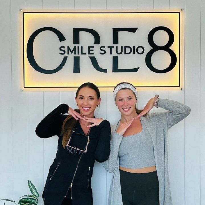 Two women posing with hand hearts in front of a lit “CARE8 Smile Studio” sign