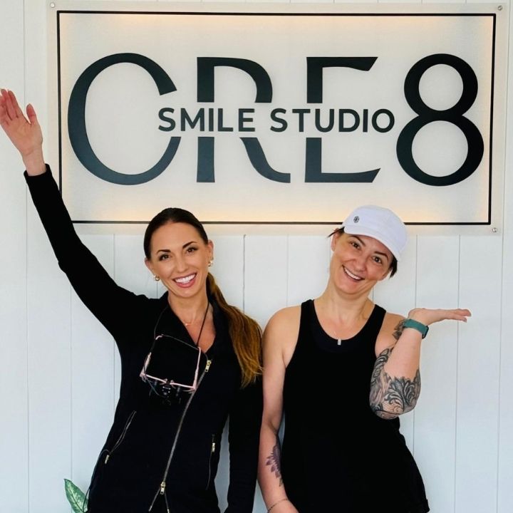 Two smiling people posing under a “Smile Studio C8” sign, one waving in a bright studio.