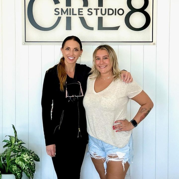 Two women standing together in front of a SMILE STUDIO sign, smiling indoors.