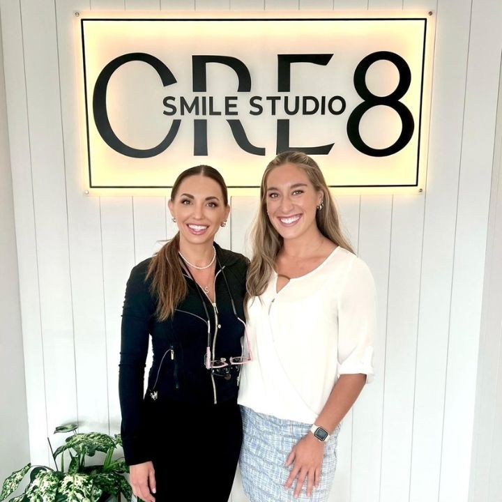 Two women smiling in front of a lit sign reading “CR8 Smile Studio”