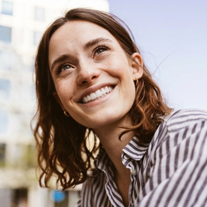 Woman with brown wavy hair looks upward, smiling. Wearing striped shirt, outdoors.