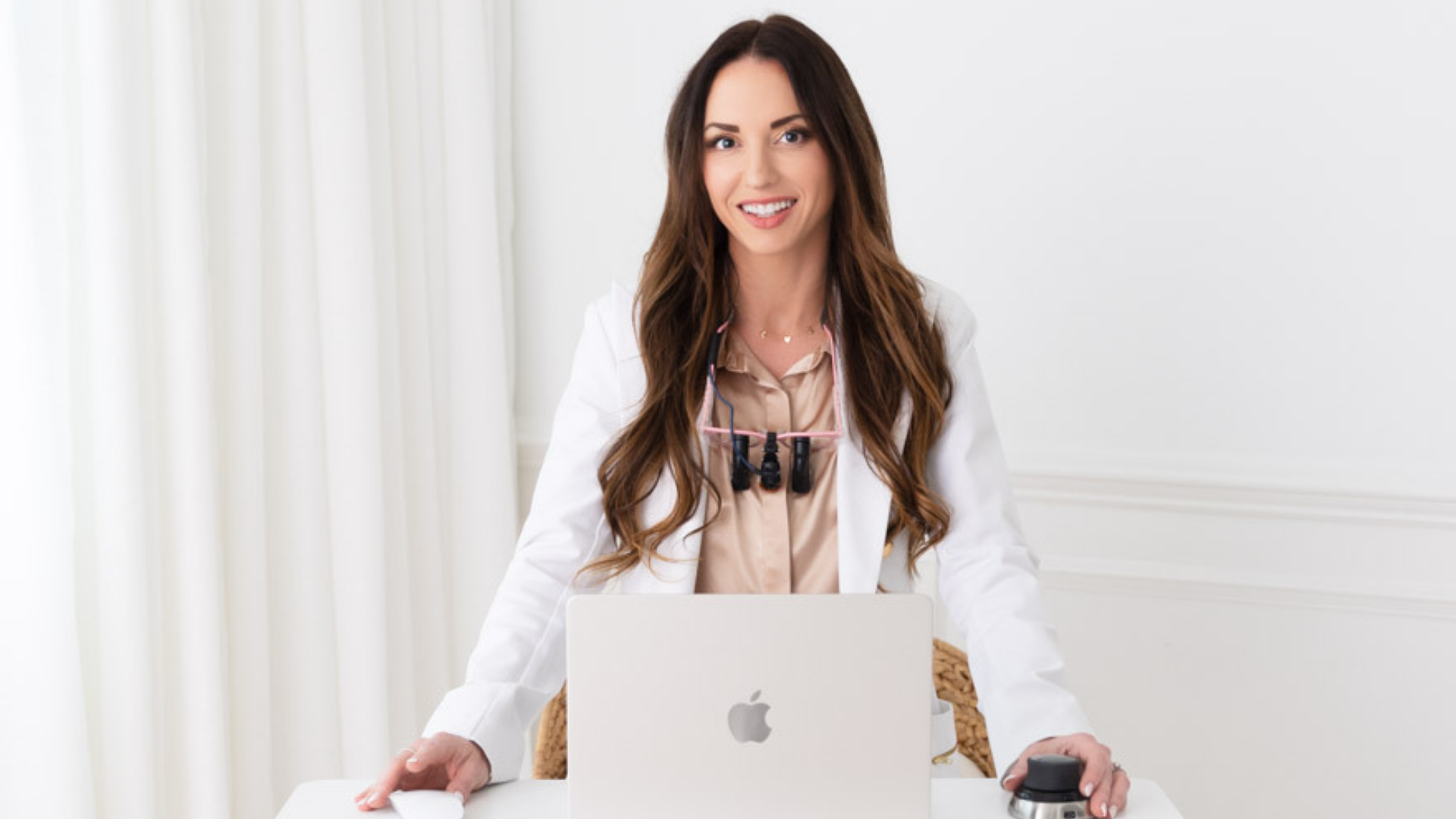 Woman smiling behind a silver laptop at a white desk in a bright office