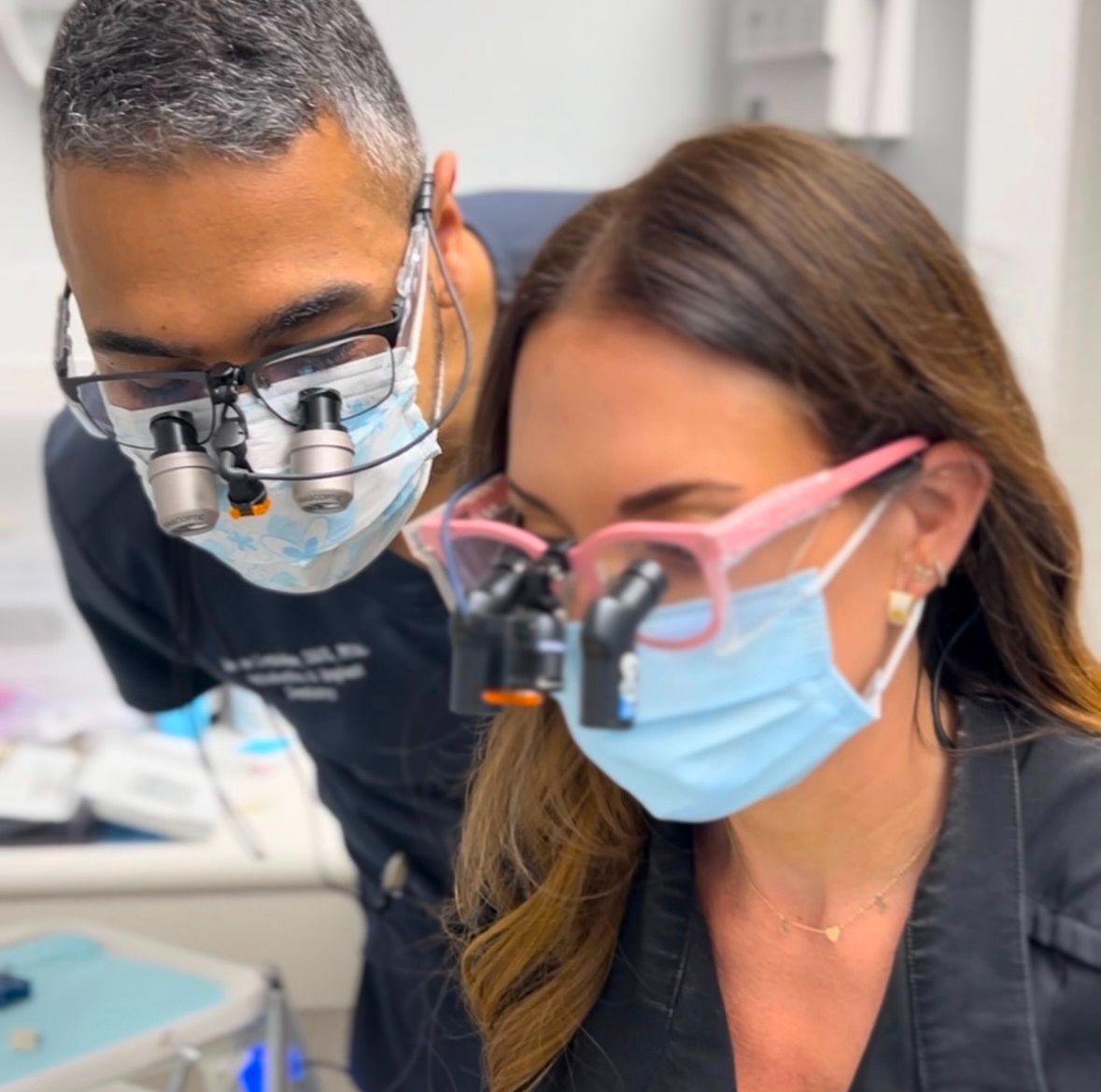 Two masked clinicians leaning over a patient during a dental procedure, wearing magnifying loupes.