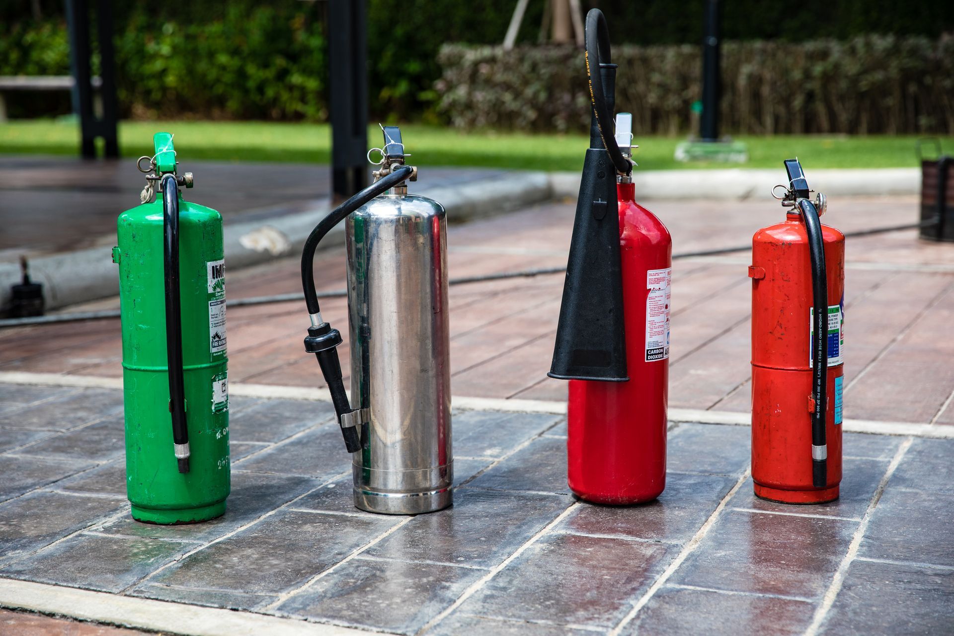 Four standing fire extinguishers in green, silver, red, and orange on a tiled pavement outdoors