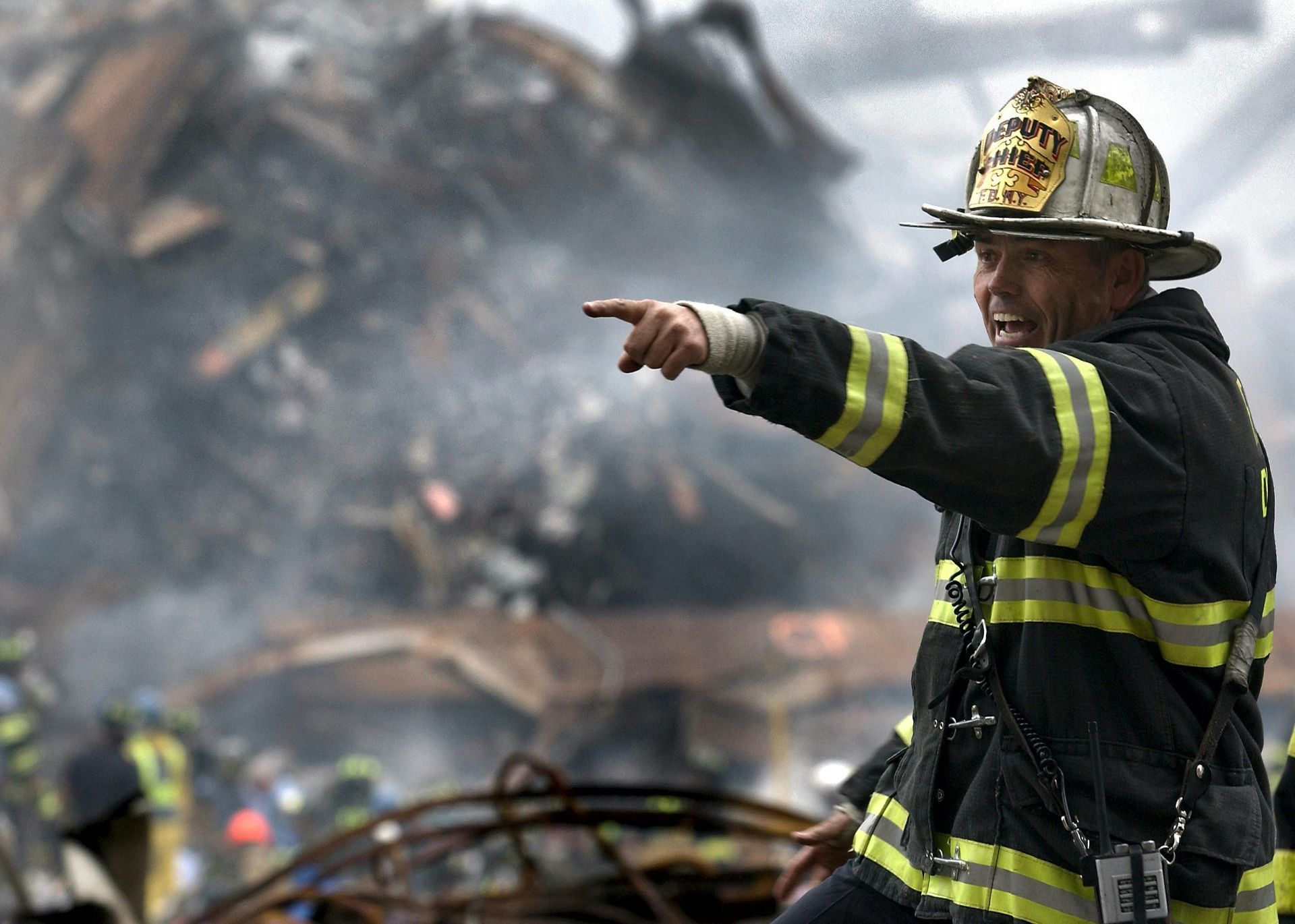 Firefighter in helmet and reflective gear points toward a smoky fire scene with burning debris.