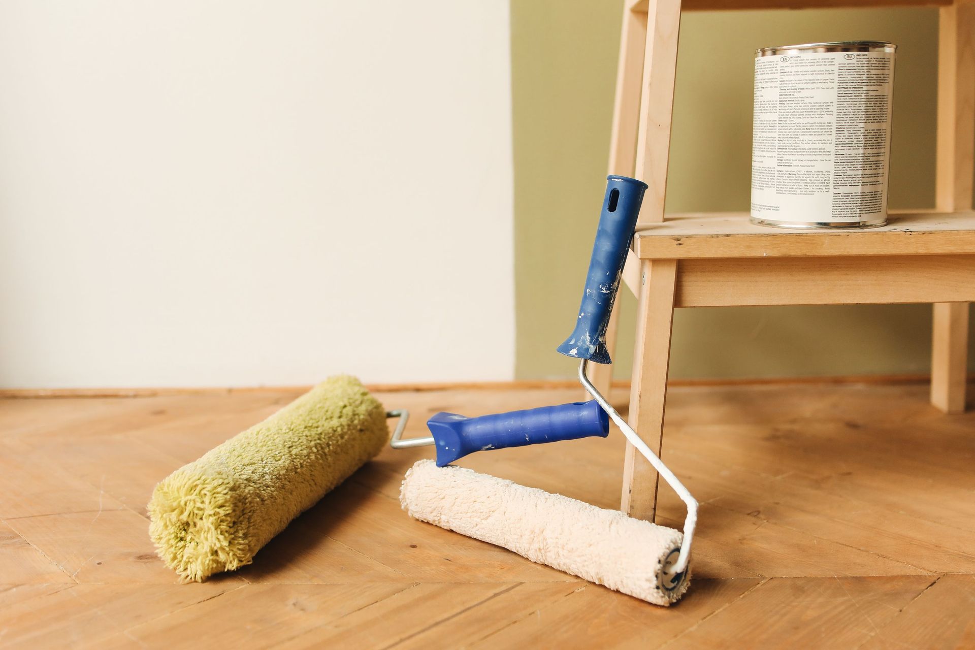 Two paint rollers and a can on a wooden floor beside a small table and wall.