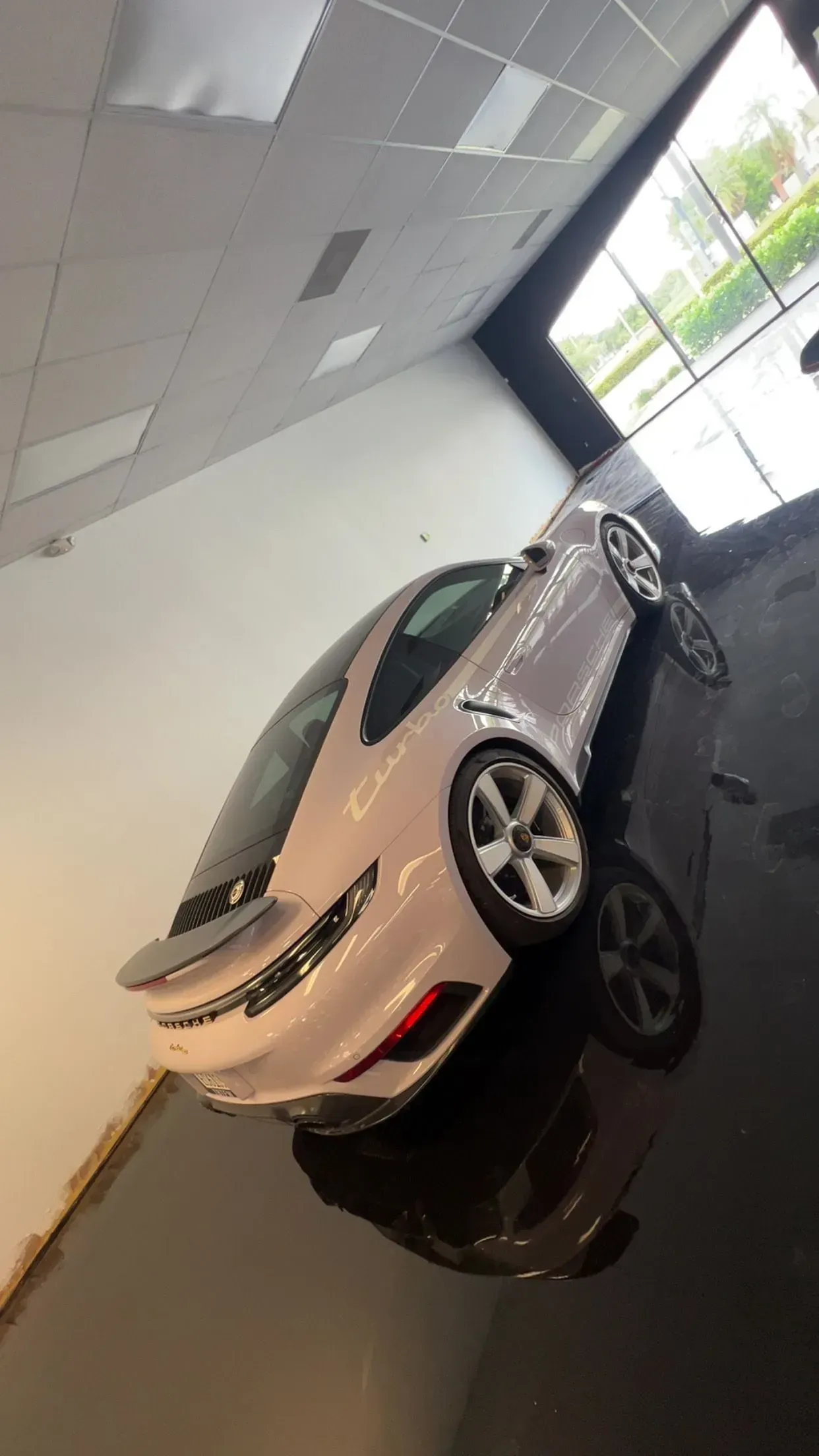 White sports car displayed indoors in a showroom, angled toward large windows.