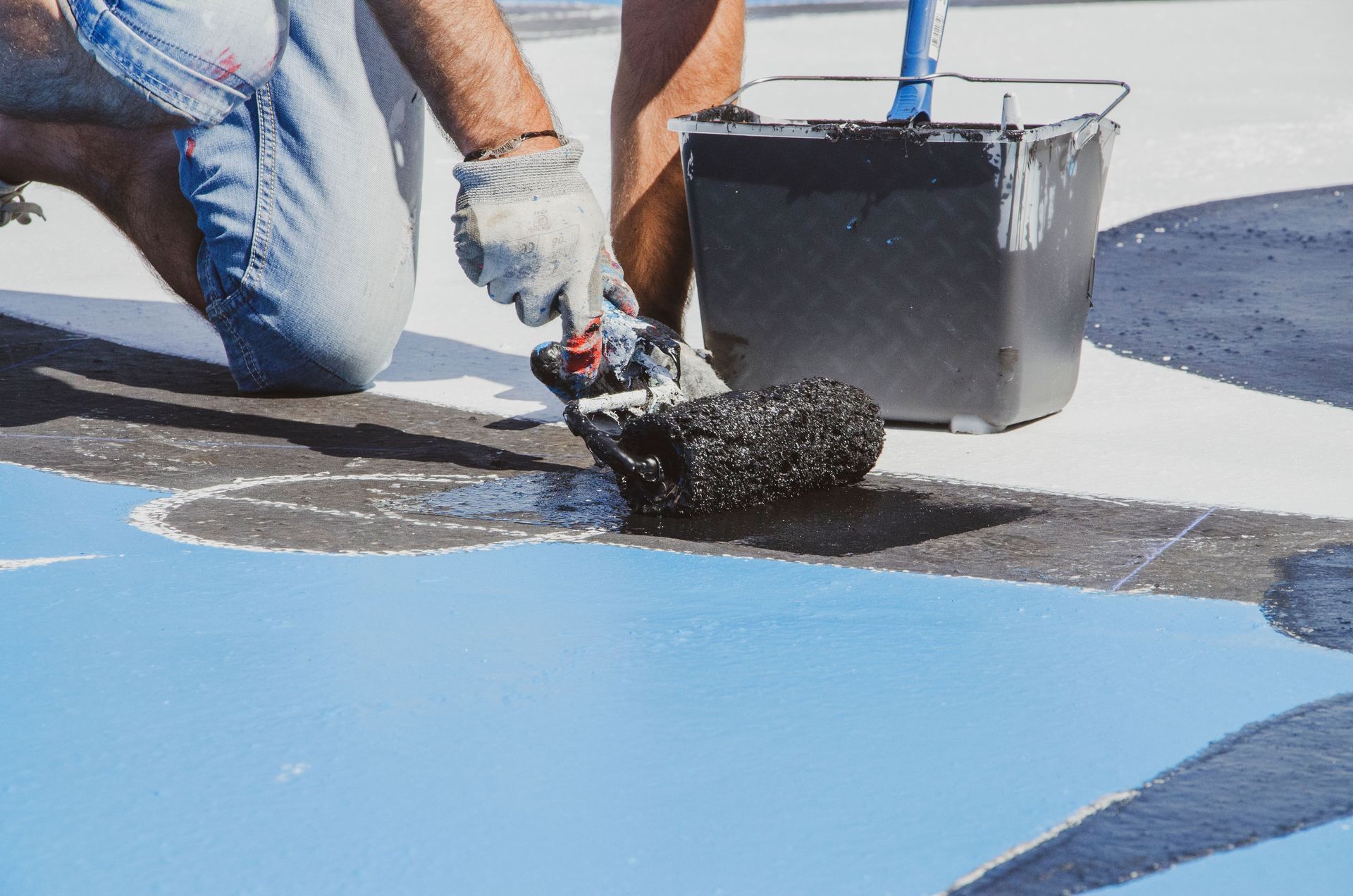 Person applying black coating with a roller on a blue rooftop surface beside a paint bucket.