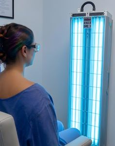 Person wearing glasses sits beside a blue UV therapy light panel in a clinic.