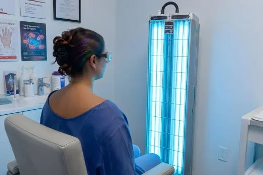 Woman seated in a clinic room using a blue light therapy panel near a desk