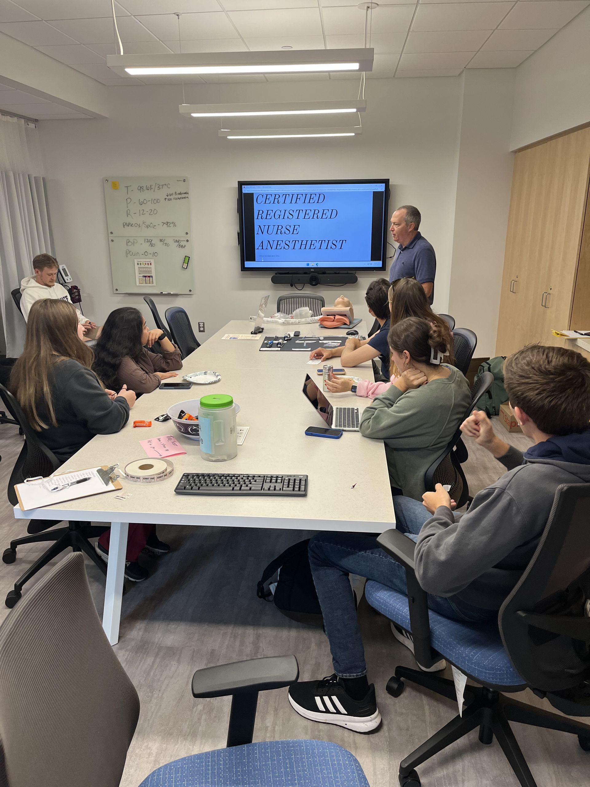 A classroom setting: teacher presents material on a screen to students seated at a large table.
