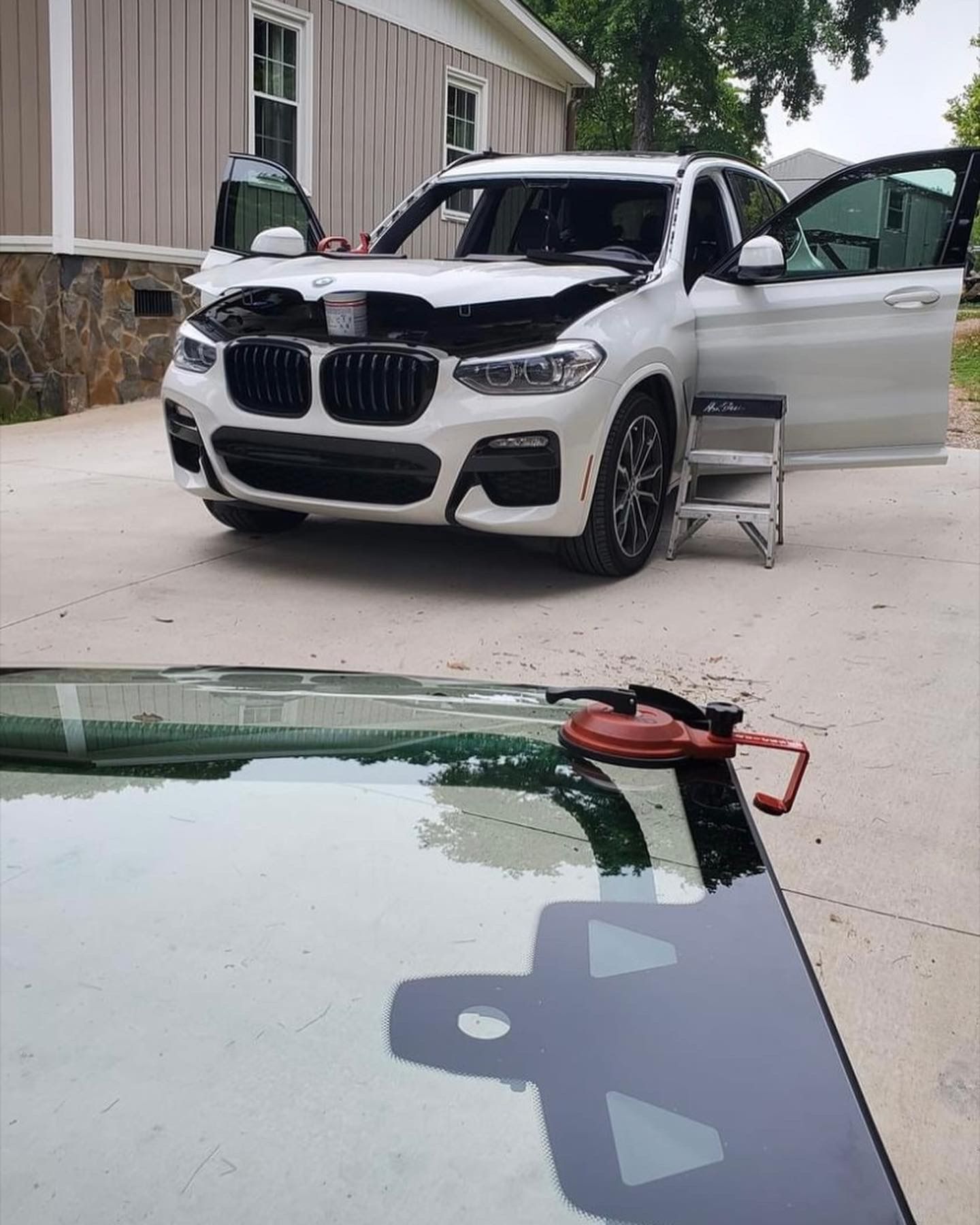 White BMW SUV with open hood and doors, windshield replacement in progress outdoors.