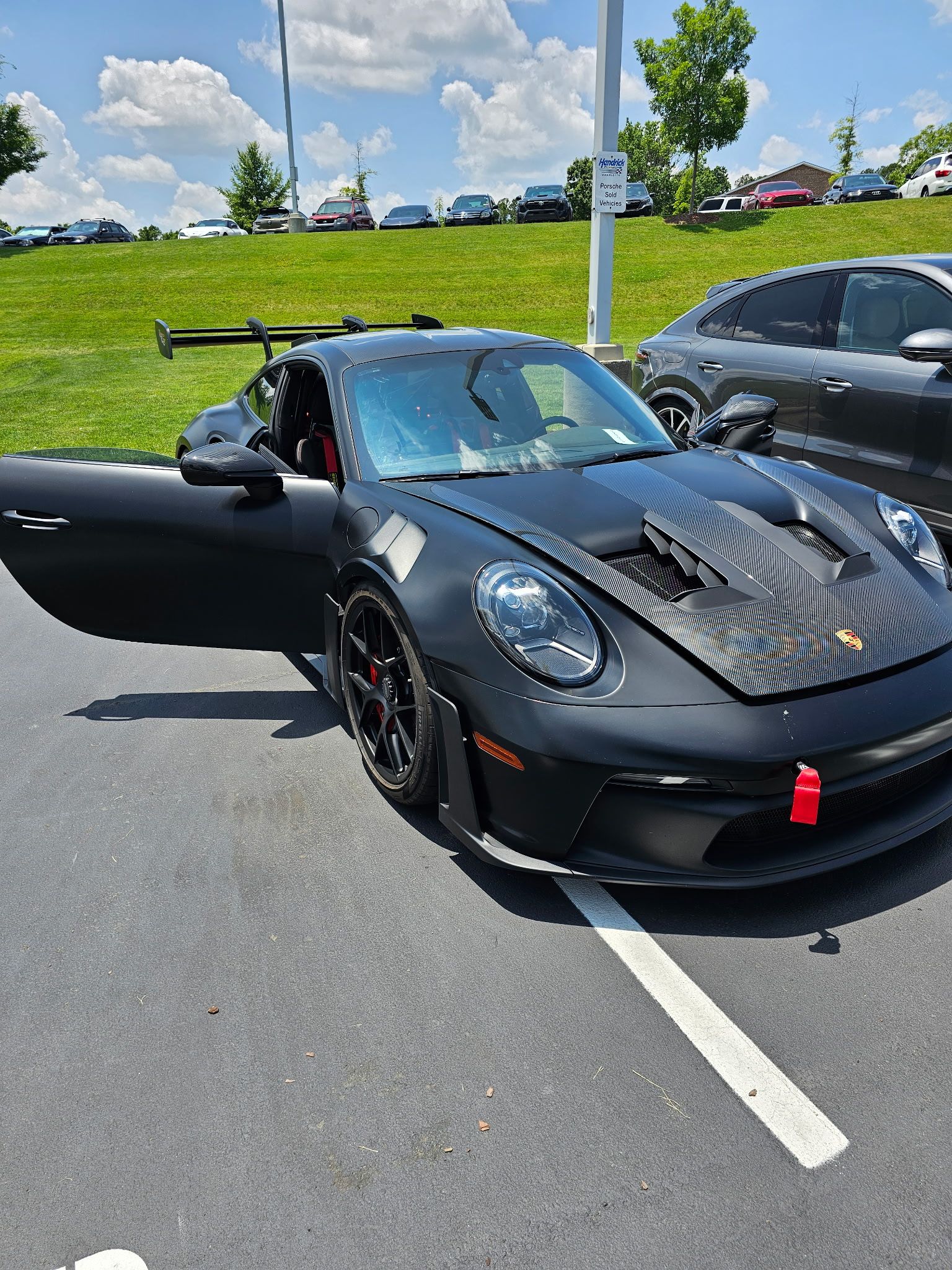 Black Porsche sports car with open door parked in a lot, carbon fiber hood, wing on back.