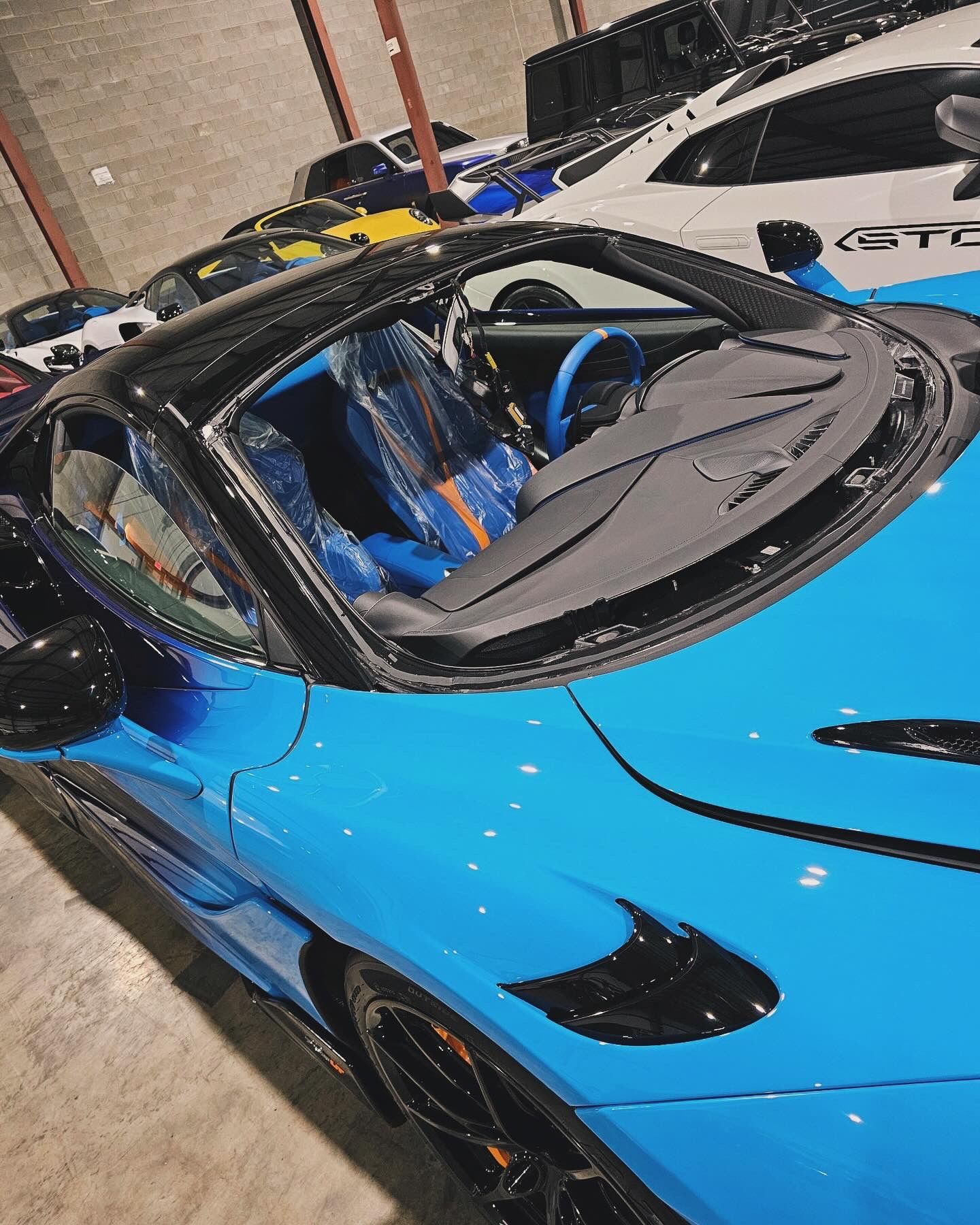Blue sports car with black accents parked inside; other cars in background.