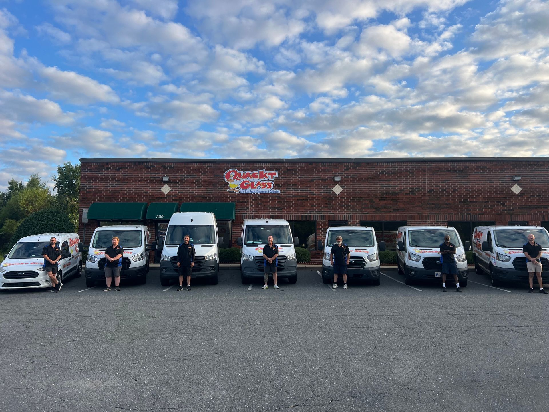 Seven service vans and staff outside a brick building under a cloudy sky. The business name is visible on the building.