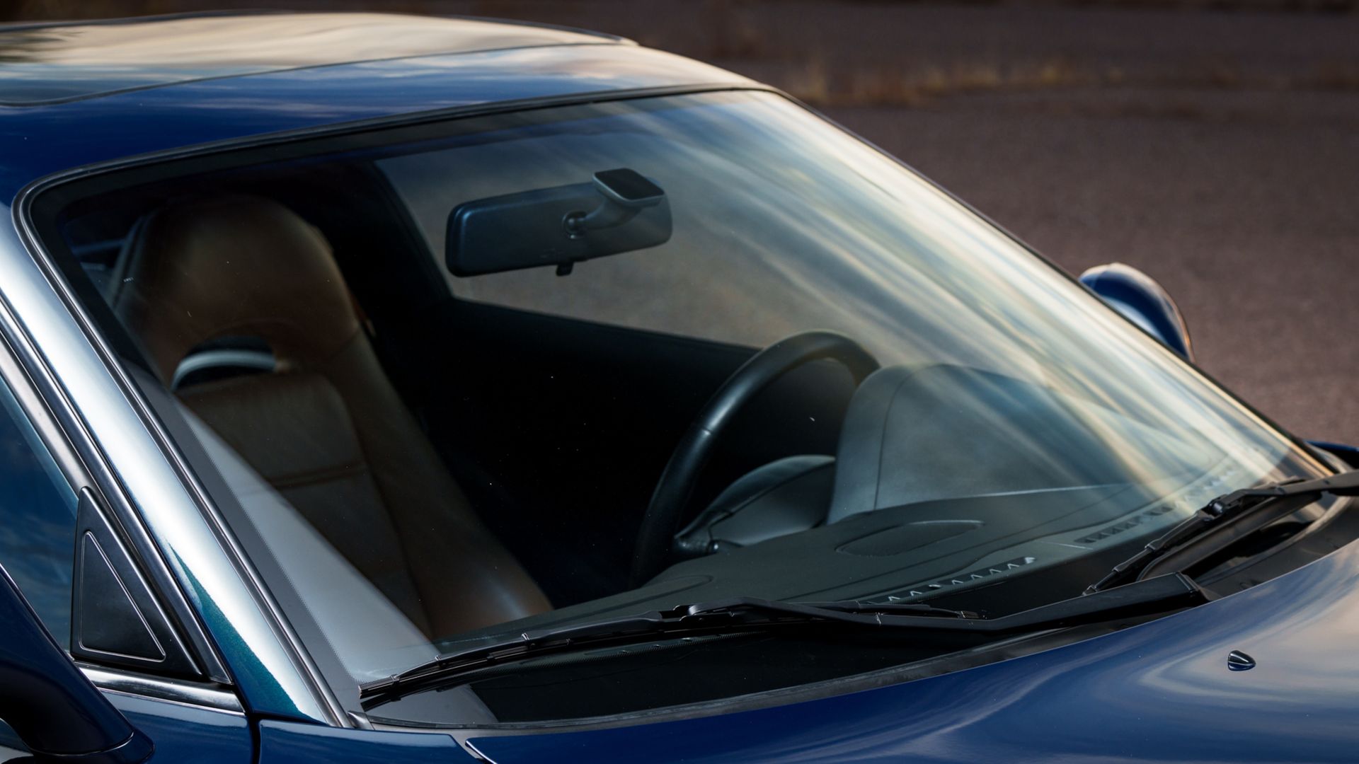 Blue convertible car interior, brown leather seat, black steering wheel, windshield with wipers, rearview mirror.