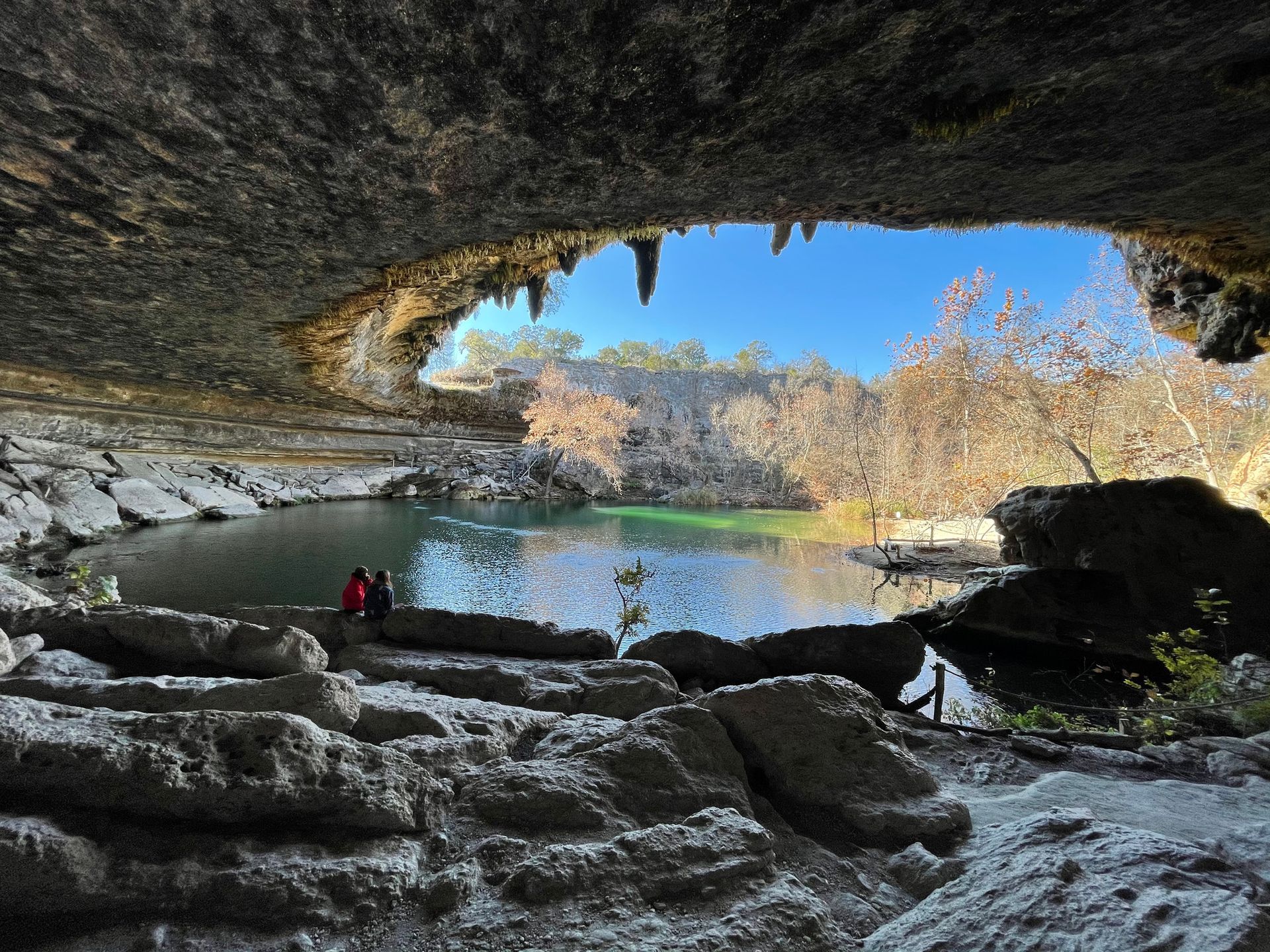 Two people sit on a rock edge looking out from a cave over a clear, turquoise swimming hole surrounded by trees.