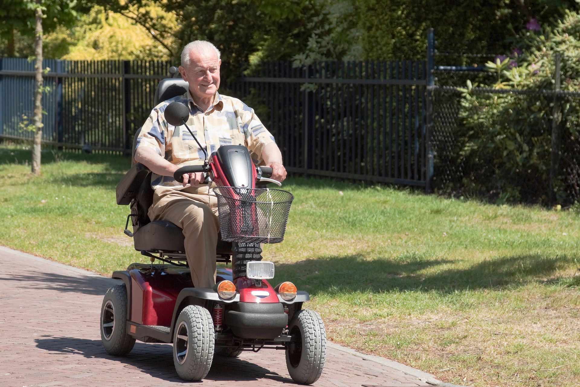 An older individual smiles while riding a red mobility scooter along a paved path next to a grassy area and fence.
