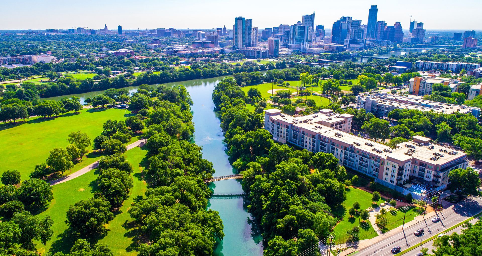 Aerial view of a river winding through a green park toward a city skyline under a clear sky.