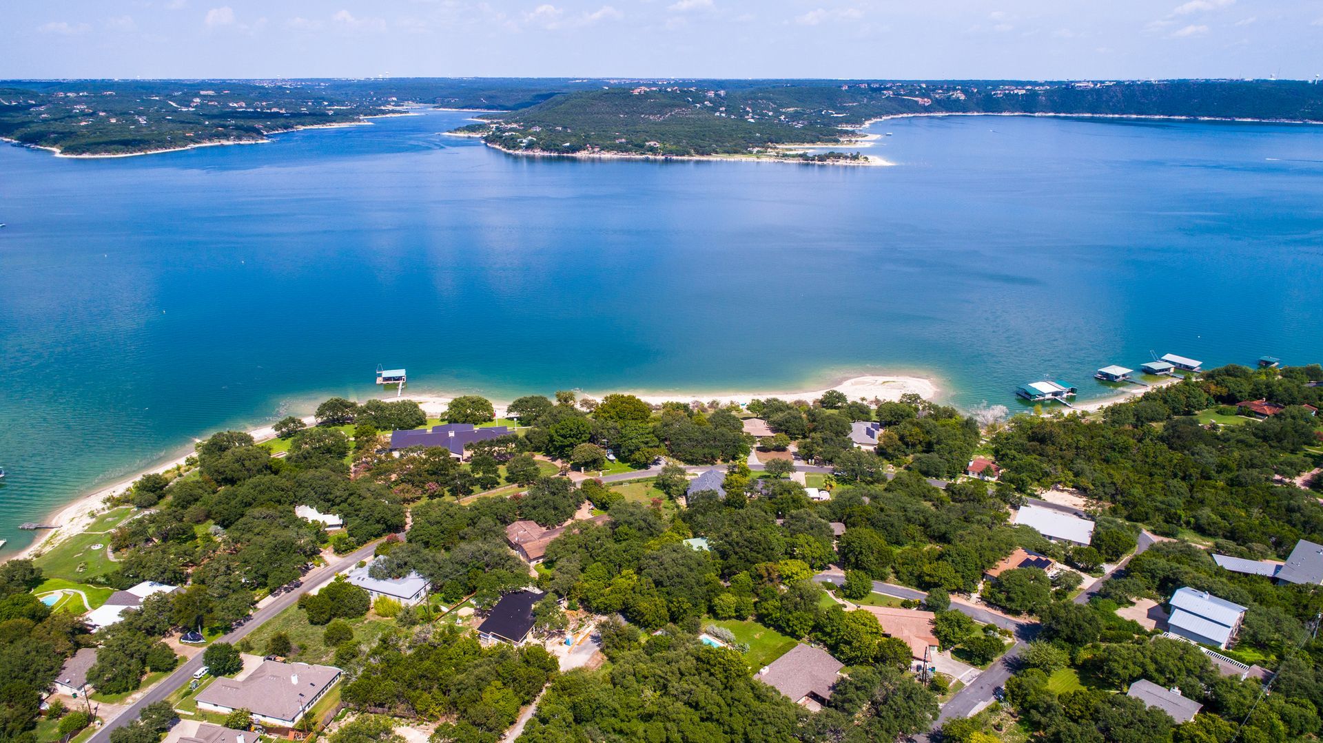 Aerial view of houses nestled in a dense, green forest along the sandy shore of a vast, blue lake on a clear day.
