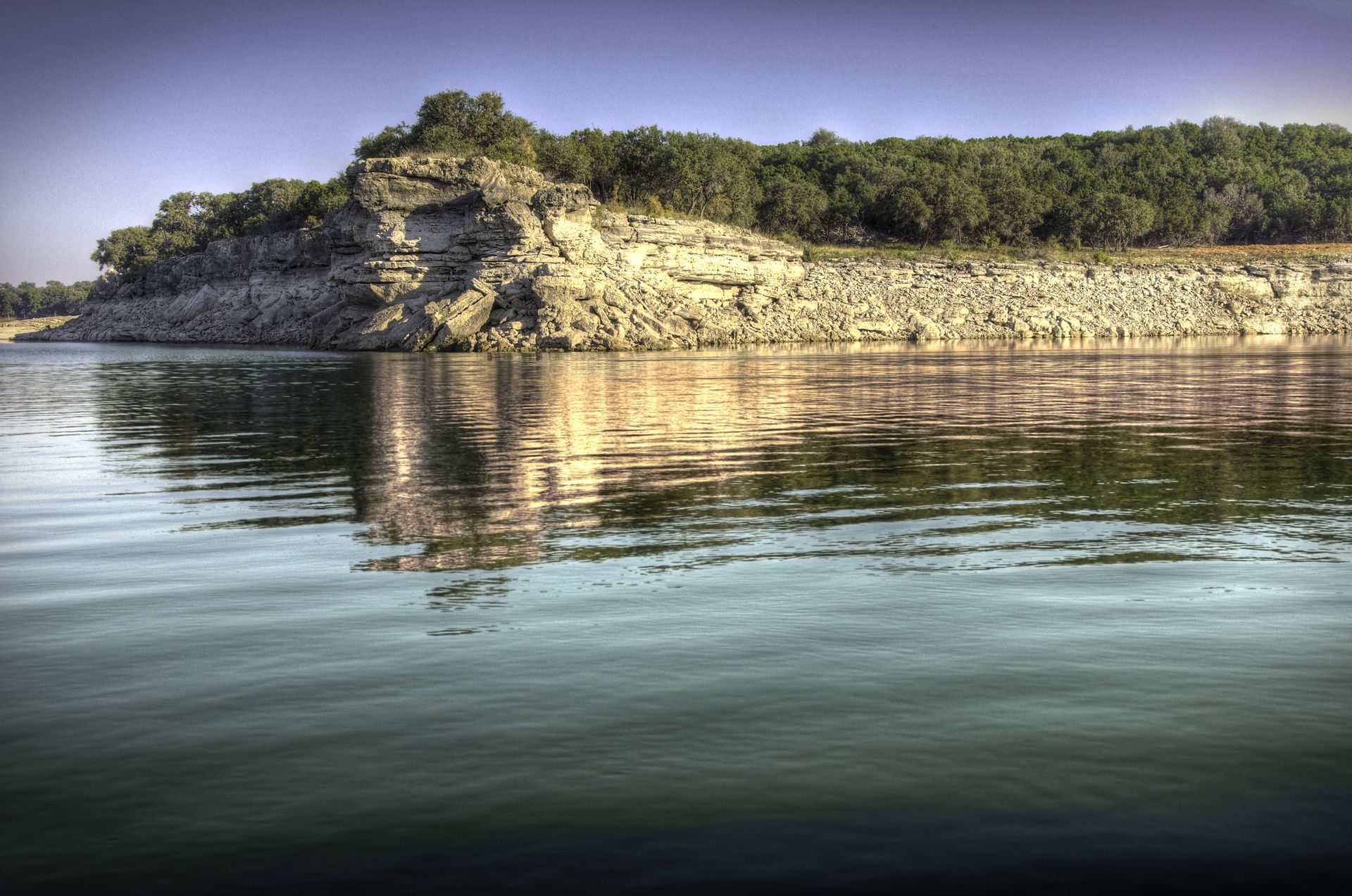 A rocky cliff with green trees overlooks a calm lake, with its reflection visible on the water's surface.
