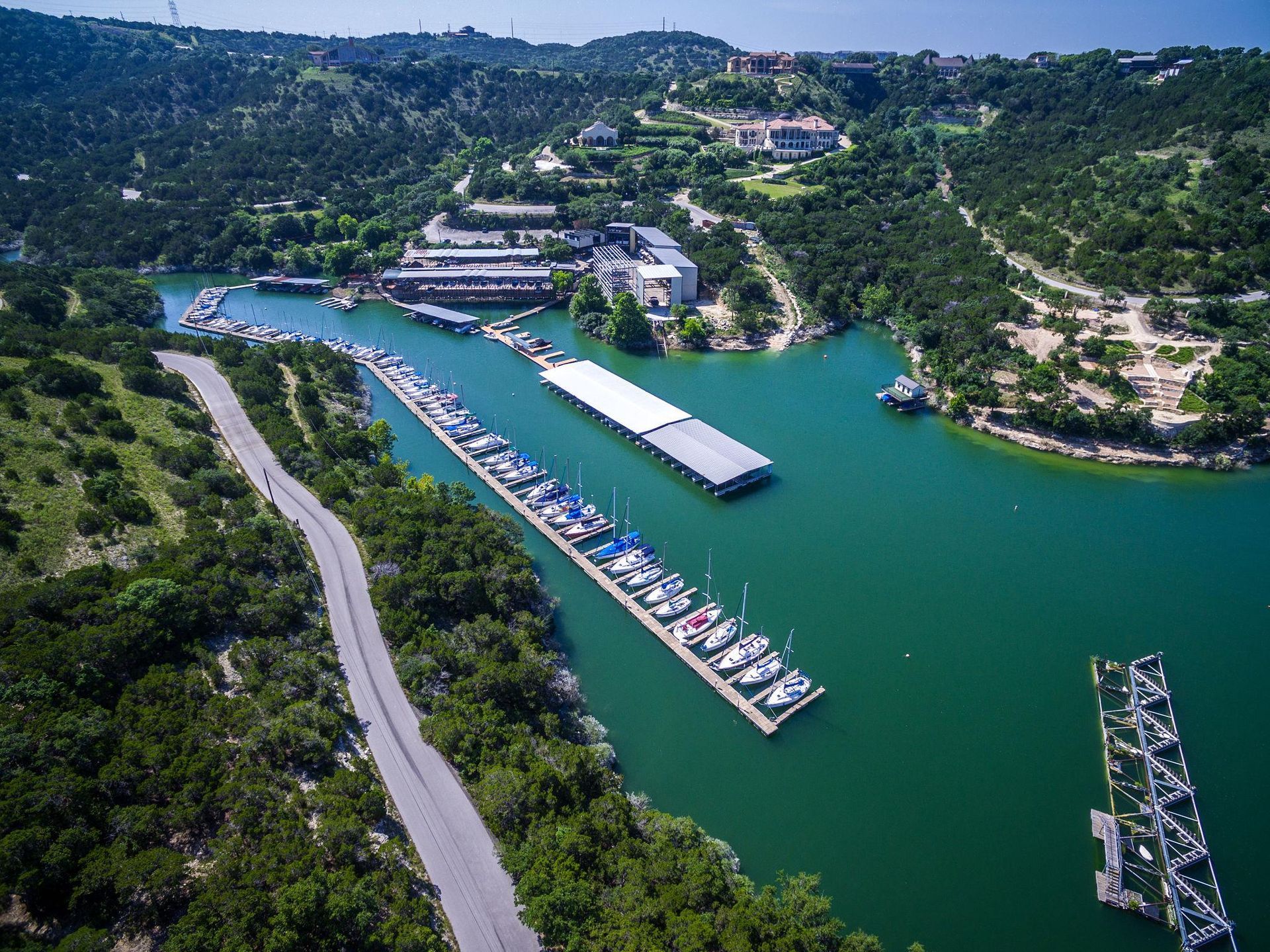 Aerial view of a marina with multiple boat docks on a calm turquoise lake, surrounded by green hills and a winding road.