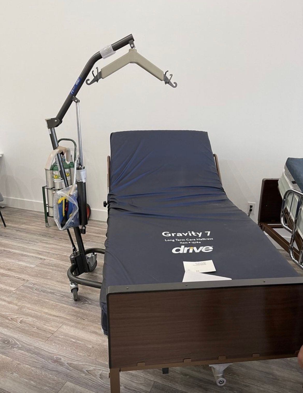 A patient lift stands beside a hospital bed with a blue Gravity 7 mattress in a room with light wood flooring.