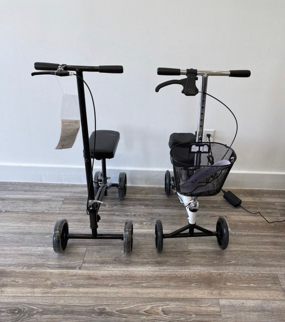 Two knee scooters standing side-by-side on a wooden floor against a white wall; one black, one white with a basket.
