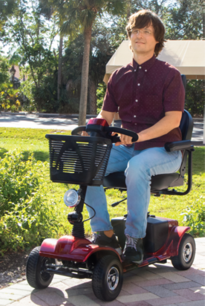 A person sits on a red mobility scooter outdoors on a paved path, holding the handlebars with a basket attached.