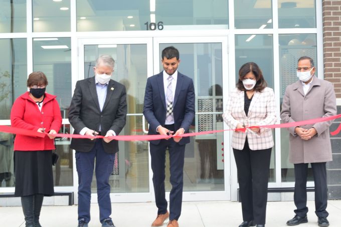 Five people stand outside a building, holding a red ribbon at a ribbon-cutting ceremony.