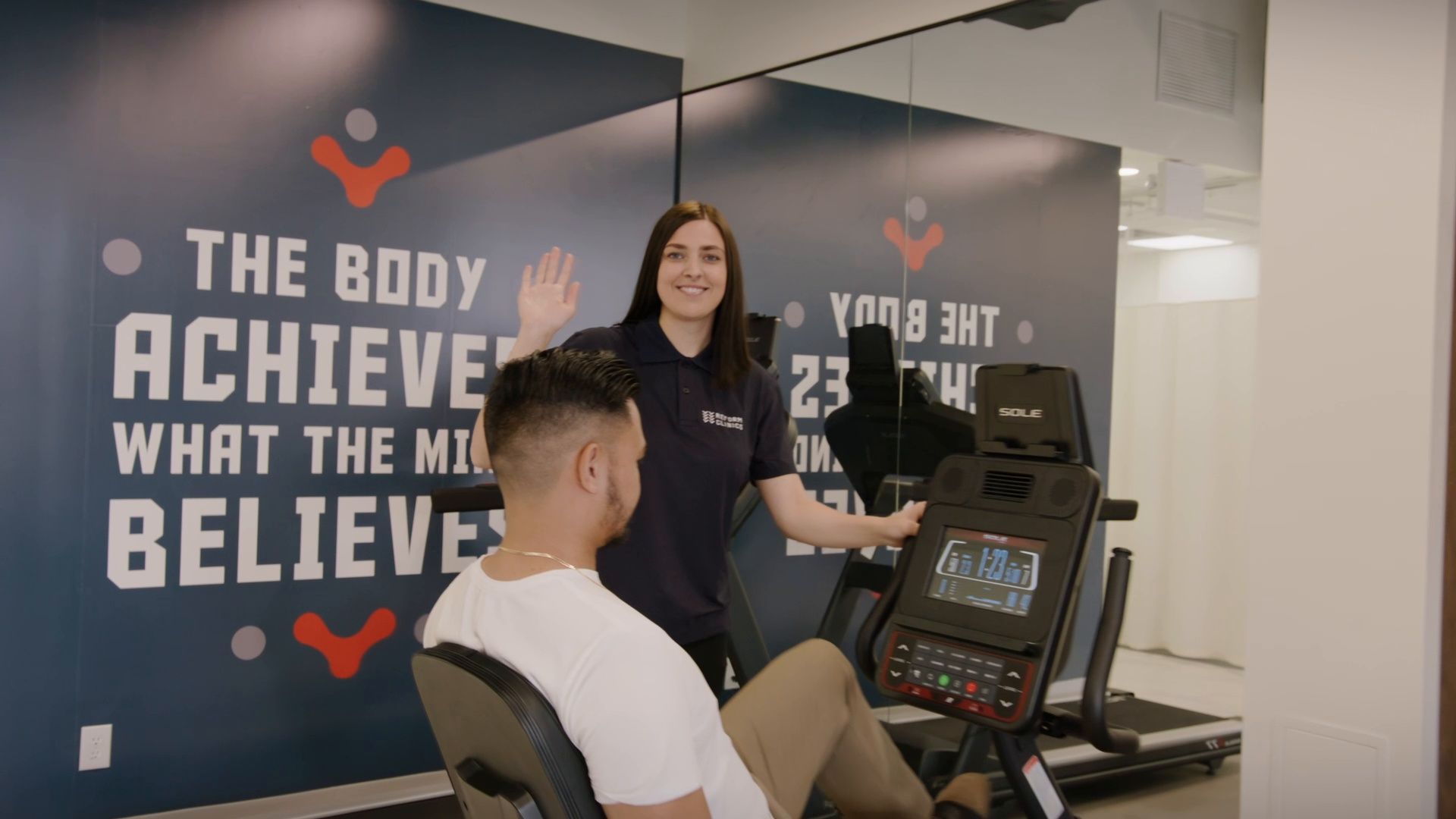 Trainer coaching a seated client at a gym, in front of a wall with motivational text.