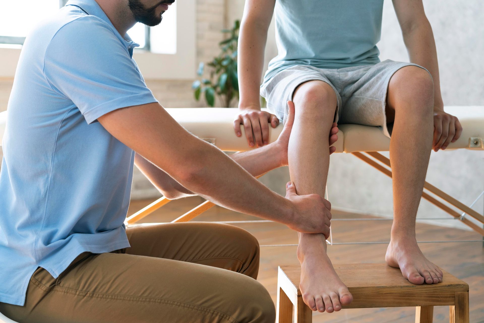 Person examining a person's leg. In a light-filled room, they sit on a stool. The person's leg rests on a wooden block.