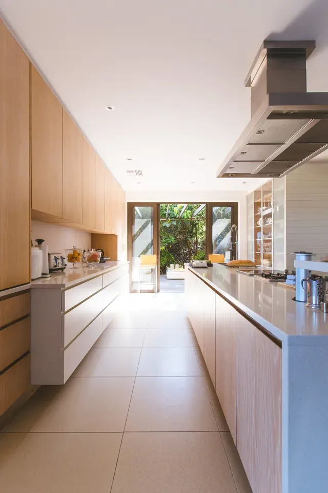 Modern kitchen with light wood cabinets, long white counter, and a view to a garden.
