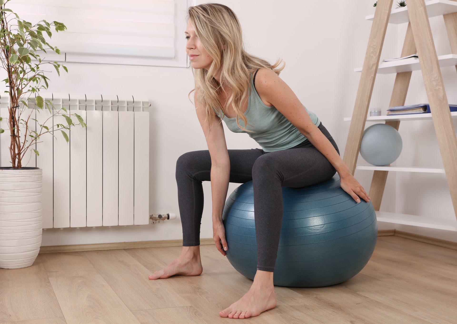 Woman sitting on a blue exercise ball in a bright room, leaning forward near a ladder shelf and radiator.