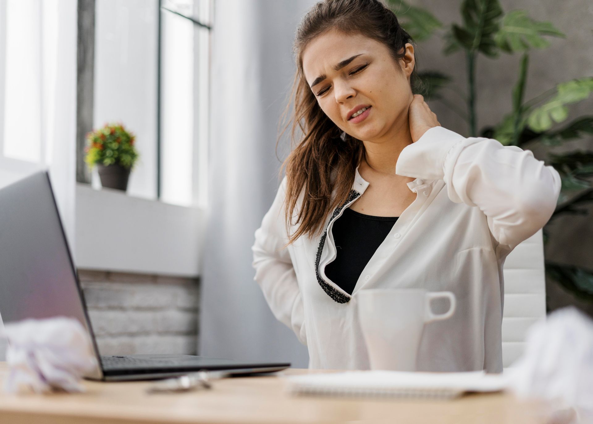a woman is sitting at a desk with a laptop and holding her neck in pain .