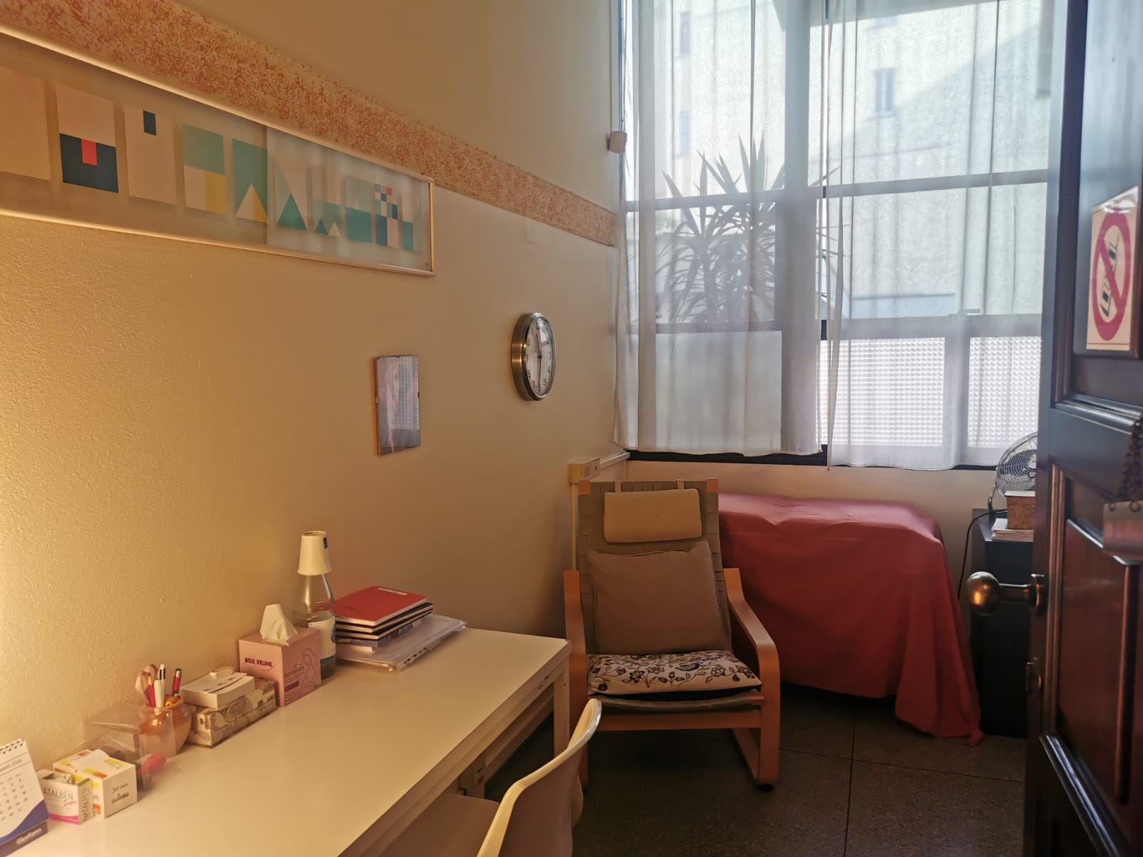 Piccola stanza con scrivania, sedia, finestra, poltrona reclinabile e coperta rossa.