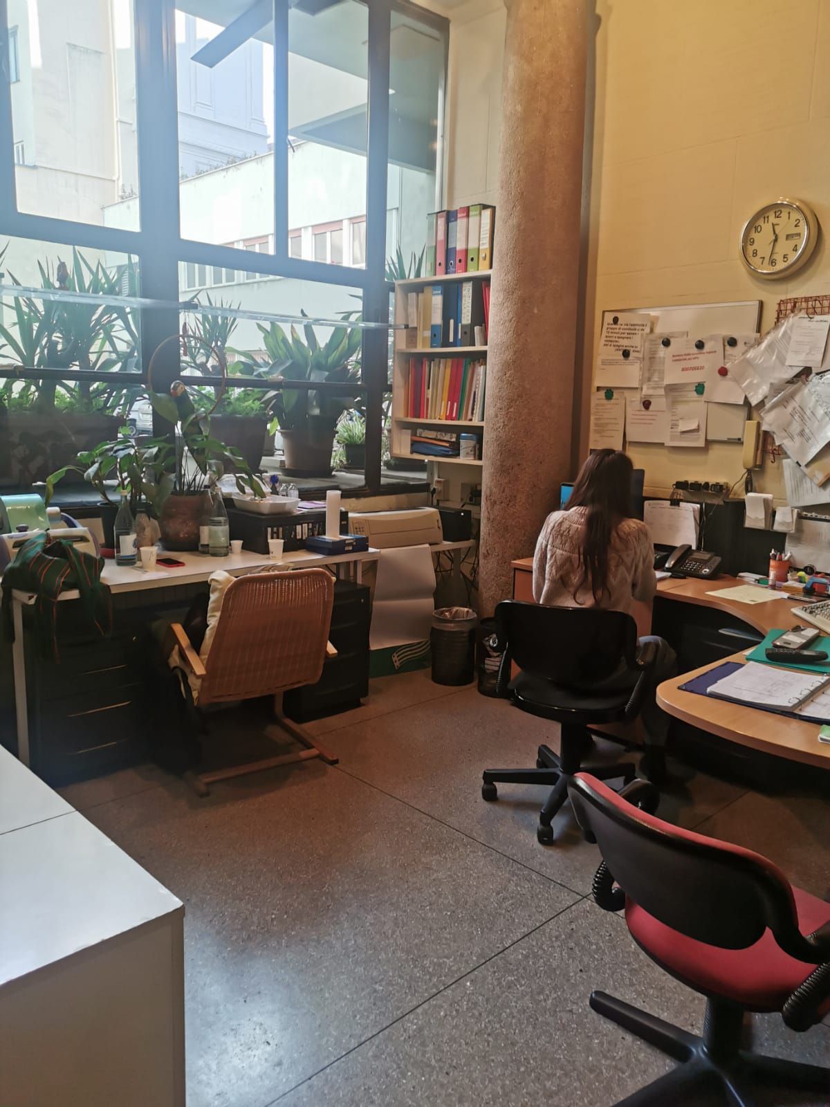 Interno di un ufficio con due scrivanie, una occupata. Finestra sullo sfondo.