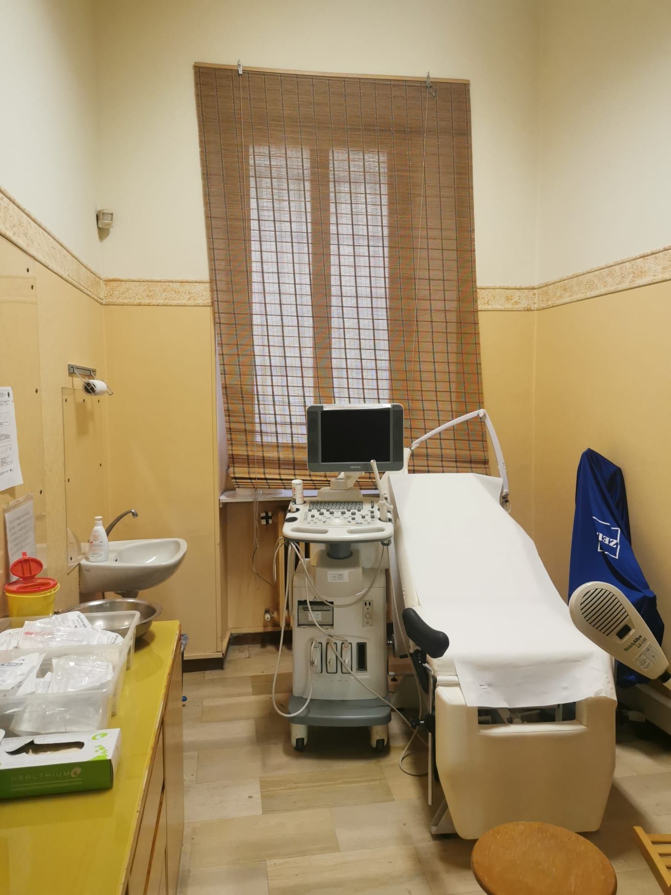 Sala visita medica: ecografo, lettino per visita, lavandino, finestra con tenda in tessuto, pareti gialle.