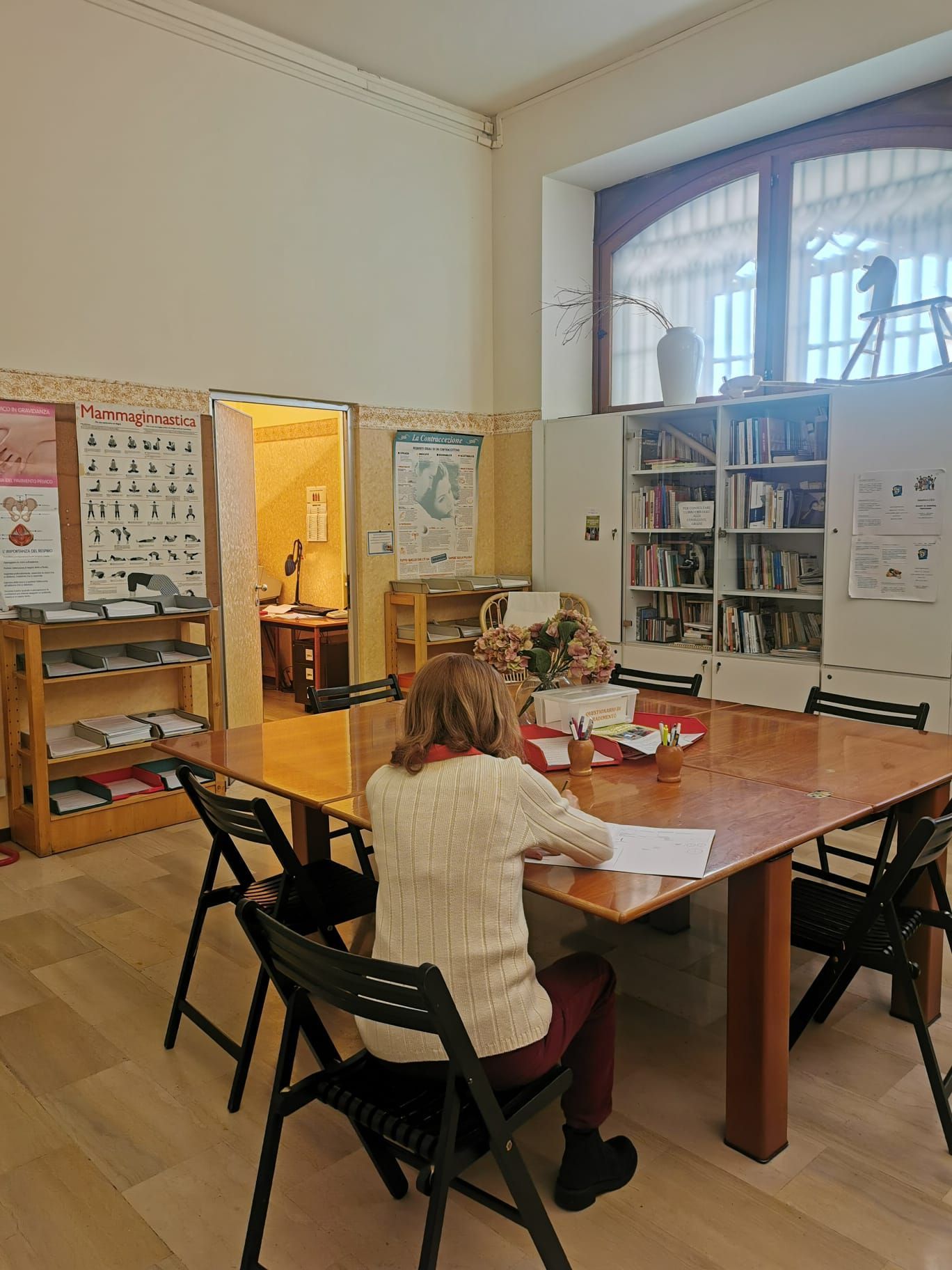 Donna seduta a un tavolo che scrive in una stanza con una libreria, una porta e delle finestre.