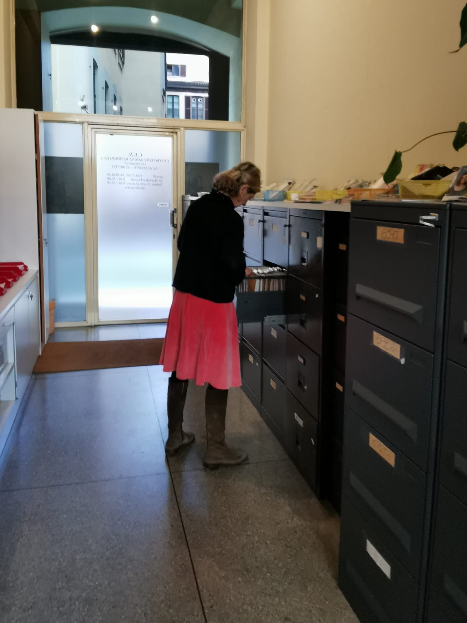 Donna che fruga nei cassetti di un archivio in un ufficio. Indossa una gonna rosa e un top nero.