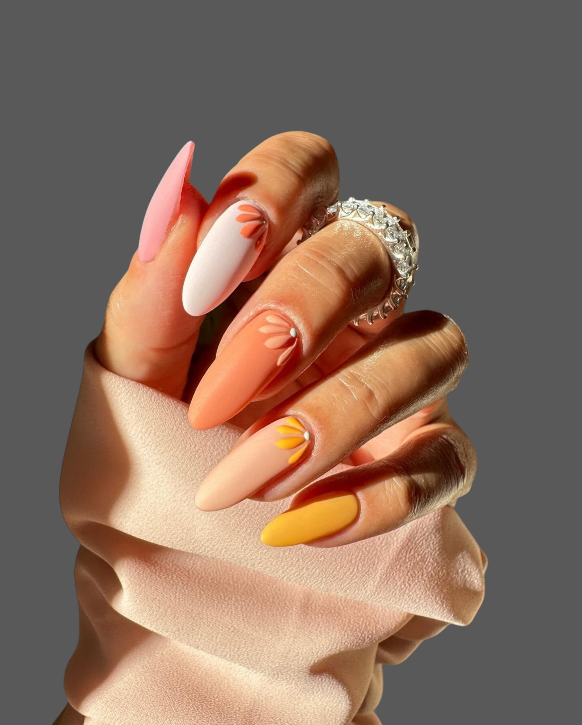 Hand with long glossy pink, peach, and yellow nails, shown in warm sunlight against a gray background